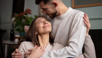 Casal aconchegante em suéteres combinando, com ela olhando para cima e sorrindo para ele, que retribui com um olhar carinhoso.