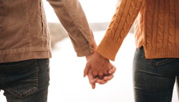 Um casal de mãos dadas, ele com uma camisa bege e ela com uma blusa de tricô laranja, simbolizando união e afeto.
