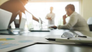 Mesa de reunião em escritório com documentos gráficos, óculos, caneta e laptops, com profissionais ao fundo desfocados.