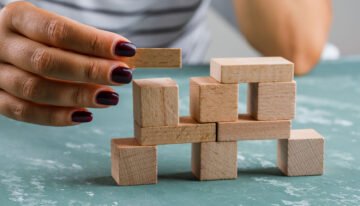Mão feminina com unhas pintadas adiciona um bloco de madeira a uma estrutura equilibrada de blocos.