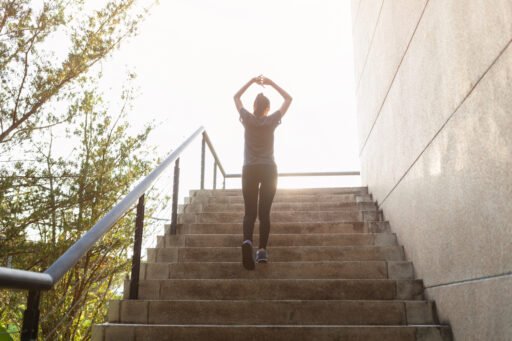 Uma mulher estica os braços acima da cabeça, celebrando a conquista ao alcançar o topo de uma escadaria ao ar livre, simbolizando como o emagrecimento pode dar a sensação de maior altura.